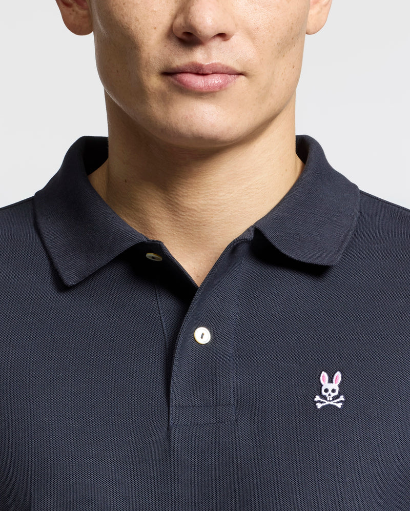 A person wears a Psycho Bunny Men's Classic Pique Polo Shirt (B6K001ARPC), made from 100% Pima cotton with mother-of-pearl buttons. The dark shirt features an embroidered pink-eared bunny logo with a skull and crossbones on the left chest against a plain white background.
