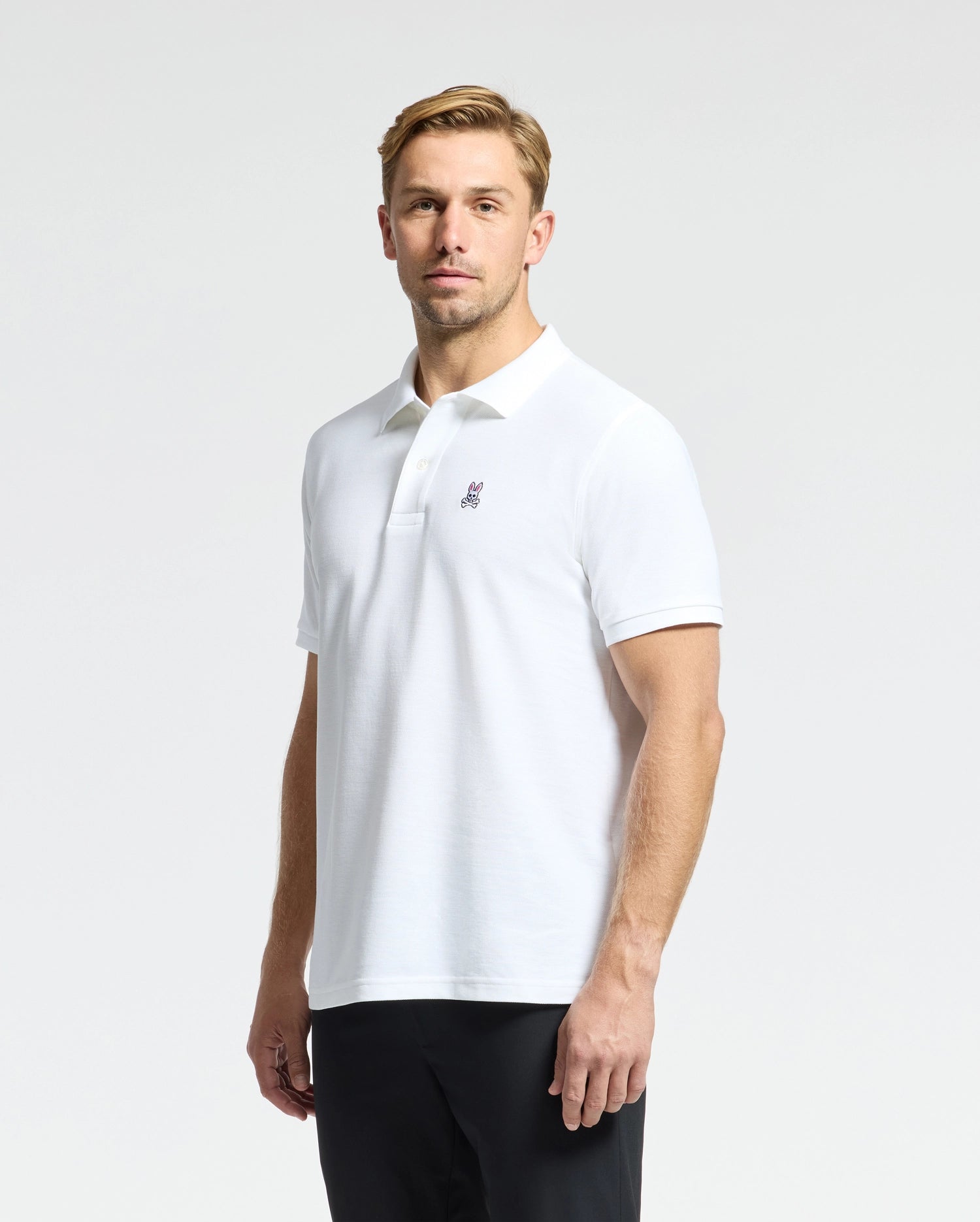 Dressed in the MENS CLASSIC PIQUE POLO SHIRT - B6K001ARPC by Psycho Bunny, featuring a small logo on the chest, a person stands against a plain background. Their white Pima cotton shirt is stylishly paired with dark pants for an effortlessly chic appearance.
