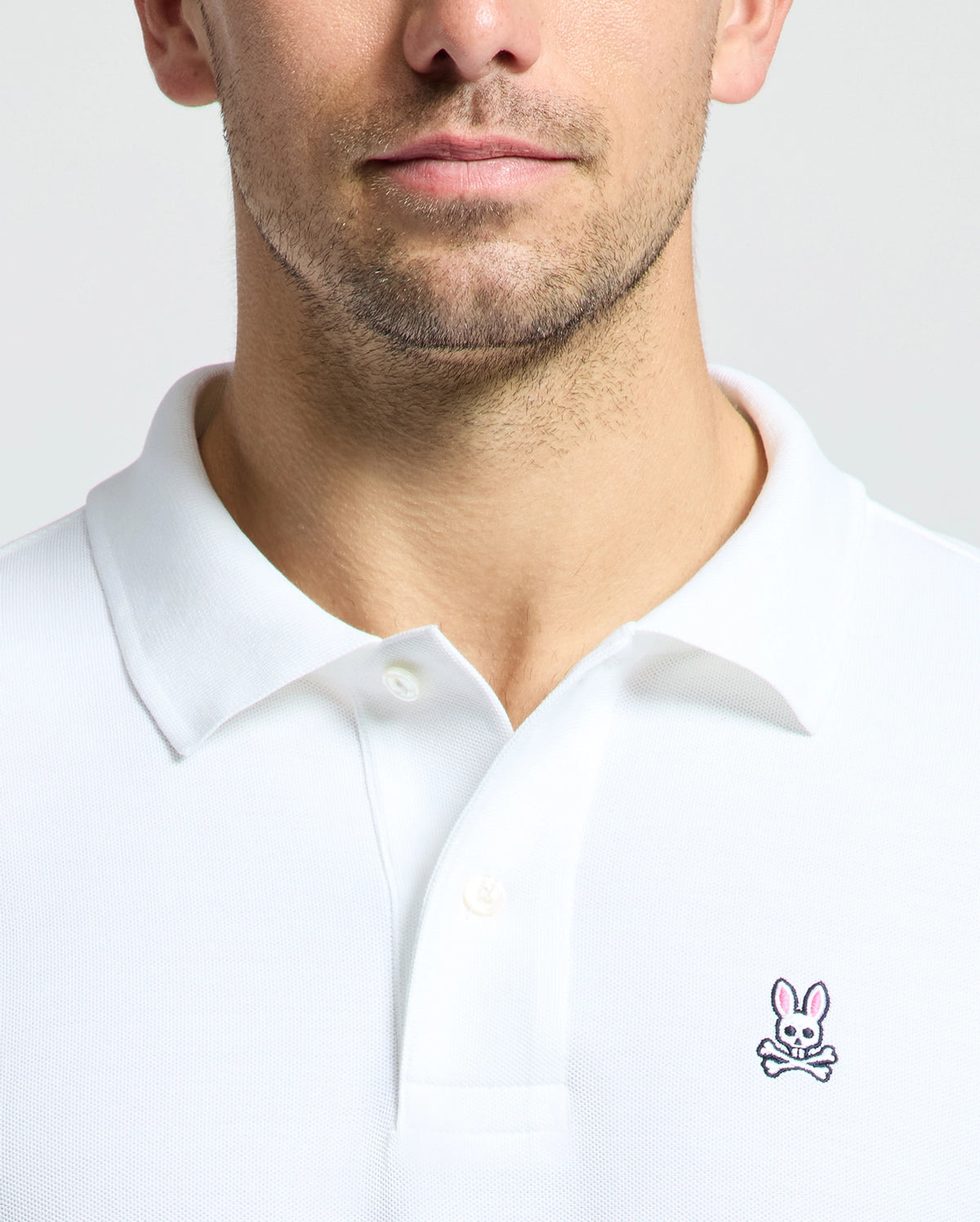 A person wears the MENS CLASSIC PIQUE POLO SHIRT - B6K001ARPC from Psycho Bunny, crafted in soft Pima cotton with the signature Psycho Bunny logo—a cartoon rabbit above crossed bones—on the left chest against a plain background. The image zooms in on the chest and collar area, highlighting the fabric's fine texture.