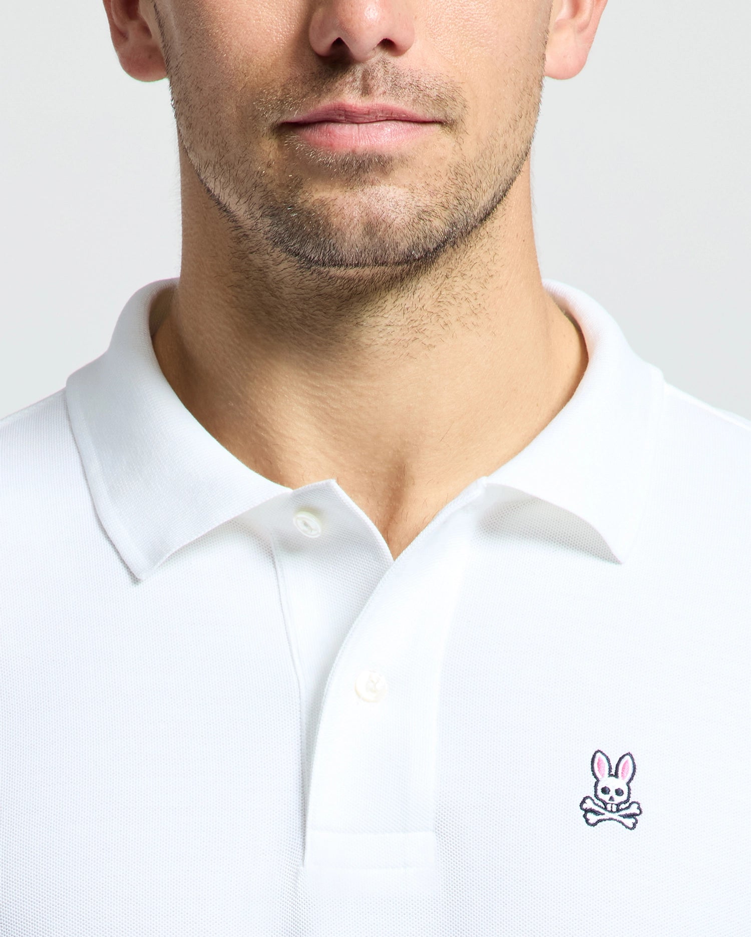 A person wears the MENS CLASSIC PIQUE POLO SHIRT - B6K001ARPC from Psycho Bunny, crafted in soft Pima cotton with the signature Psycho Bunny logo—a cartoon rabbit above crossed bones—on the left chest against a plain background. The image zooms in on the chest and collar area, highlighting the fabric's fine texture.