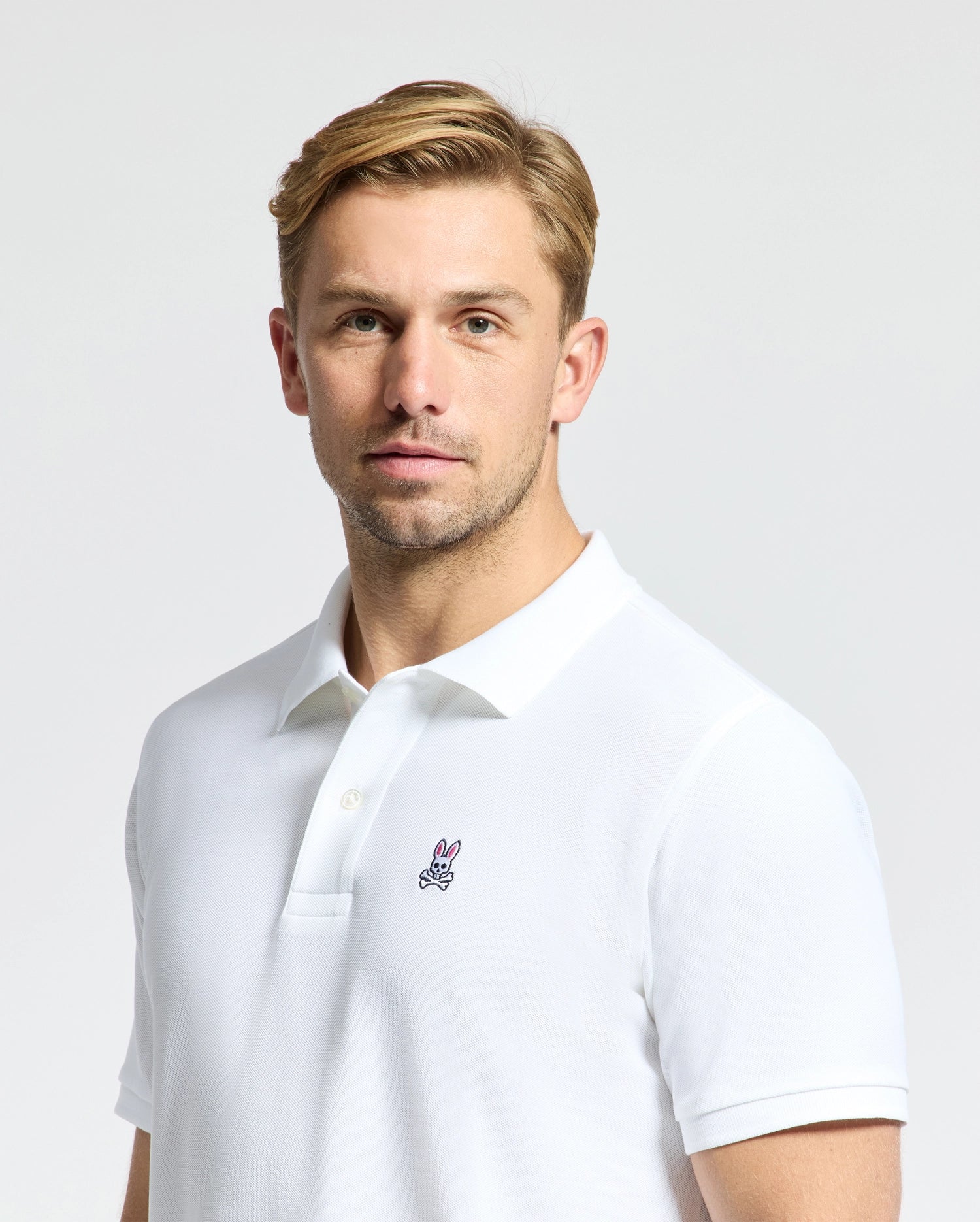 A person with short, light-colored hair is wearing the Psycho Bunny Men's Classic Pique Polo Shirt (B6K001ARPC), crafted from soft Pima cotton and featuring the signature embroidered Psycho Bunny logo on the chest. The setting has a plain, light-colored background.