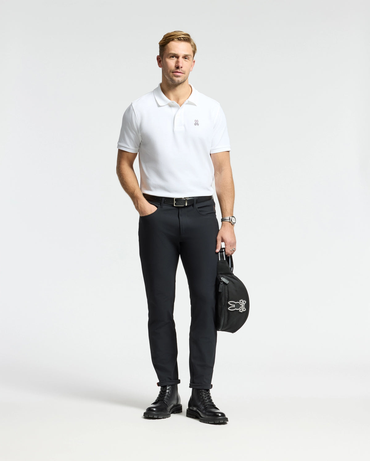 A man dressed in a MENS CLASSIC PIQUE POLO SHIRT - B6K001ARPC by Psycho Bunny stands against a plain background, paired with black pants and leather boots. In his left hand, he holds a black bag adorned with the Psycho Bunny logo, and an elegant watch adds understated sophistication to his look.