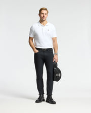 A man dressed in a MENS CLASSIC PIQUE POLO SHIRT - B6K001ARPC by Psycho Bunny stands against a plain background, paired with black pants and leather boots. In his left hand, he holds a black bag adorned with the Psycho Bunny logo, and an elegant watch adds understated sophistication to his look.