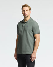 A person wears a forest green Psycho Bunny Men's Classic Piqué Polo, identified by product code B6K001B200, featuring an embroidered Bunny logo on the chest. The Pima cotton fabric ensures comfort as they pair it with black pants against a plain white background.