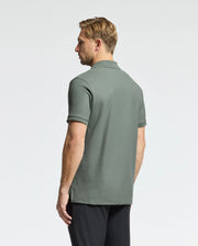 A man with short blond hair faces away in a studio, wearing Psycho Bunny's green Men's Classic Piqué Polo (B6K001B200) with an embroidered Bunny logo and black pants. The light-colored backdrop enhances the subtle elegance of its Pima cotton fabric.