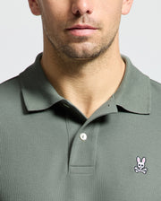 A close-up shows a man's torso wearing a Psycho Bunny Men's Classic Pique Polo Shirt, B6K001B200, in olive green. Made from soft Pima cotton, it features an embroidered Bunny logo with crossbones on the chest. The background is neutral and light.