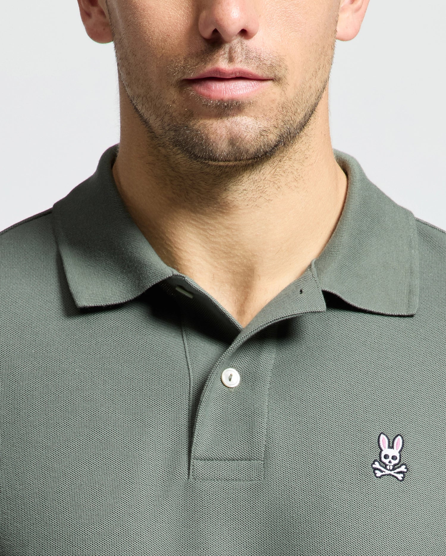 A close-up shows a man's torso wearing a Psycho Bunny Men's Classic Pique Polo Shirt, B6K001B200, in olive green. Made from soft Pima cotton, it features an embroidered Bunny logo with crossbones on the chest. The background is neutral and light.