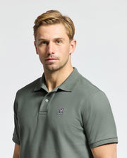 A person with short blonde hair is wearing a Psycho Bunny MENS CLASSIC PIQUE POLO SHIRT - B6K001B200 made from Pima cotton, with an embroidered Bunny logo on the left chest against a plain light gray background.