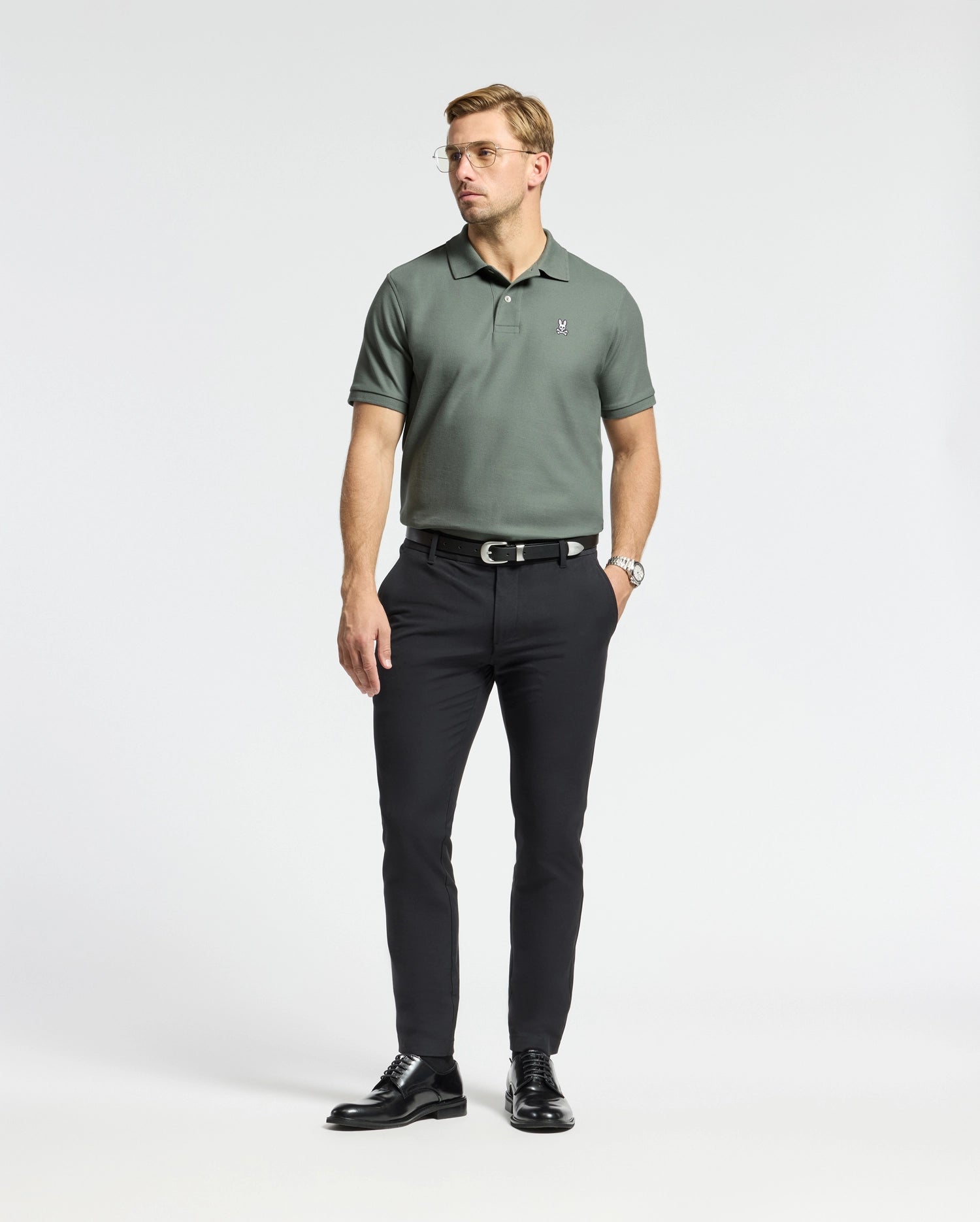 A man stands against a plain white background, wearing glasses, black pants, and shiny black shoes. He wears a Psycho Bunny men's classic Pima cotton polo in green (B6K001B200), with an embroidered Bunny logo visible. His arms rest by his sides as he looks slightly to his right.