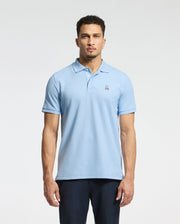 A man stands facing forward, wearing a Psycho Bunny Men's Classic Pique Polo Shirt in light blue, featuring soft Pima cotton and an embroidered Bunny logo on the chest, paired with dark pants against a plain white background.