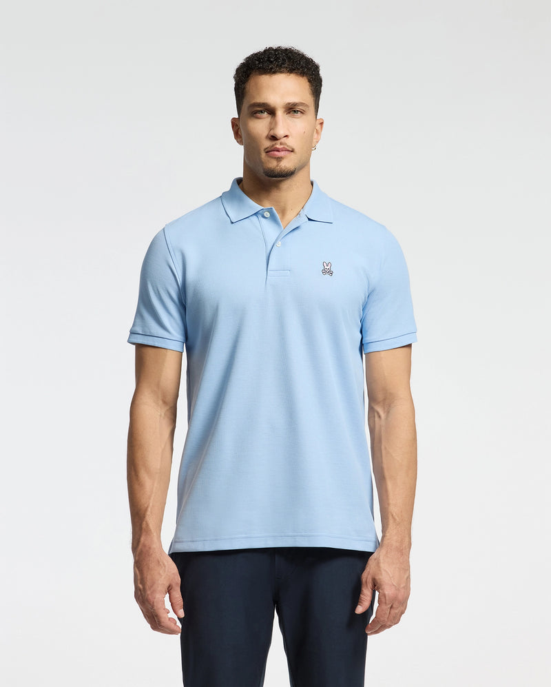 A man stands facing forward, wearing a Psycho Bunny Men's Classic Pique Polo Shirt in light blue, featuring soft Pima cotton and an embroidered Bunny logo on the chest, paired with dark pants against a plain white background.