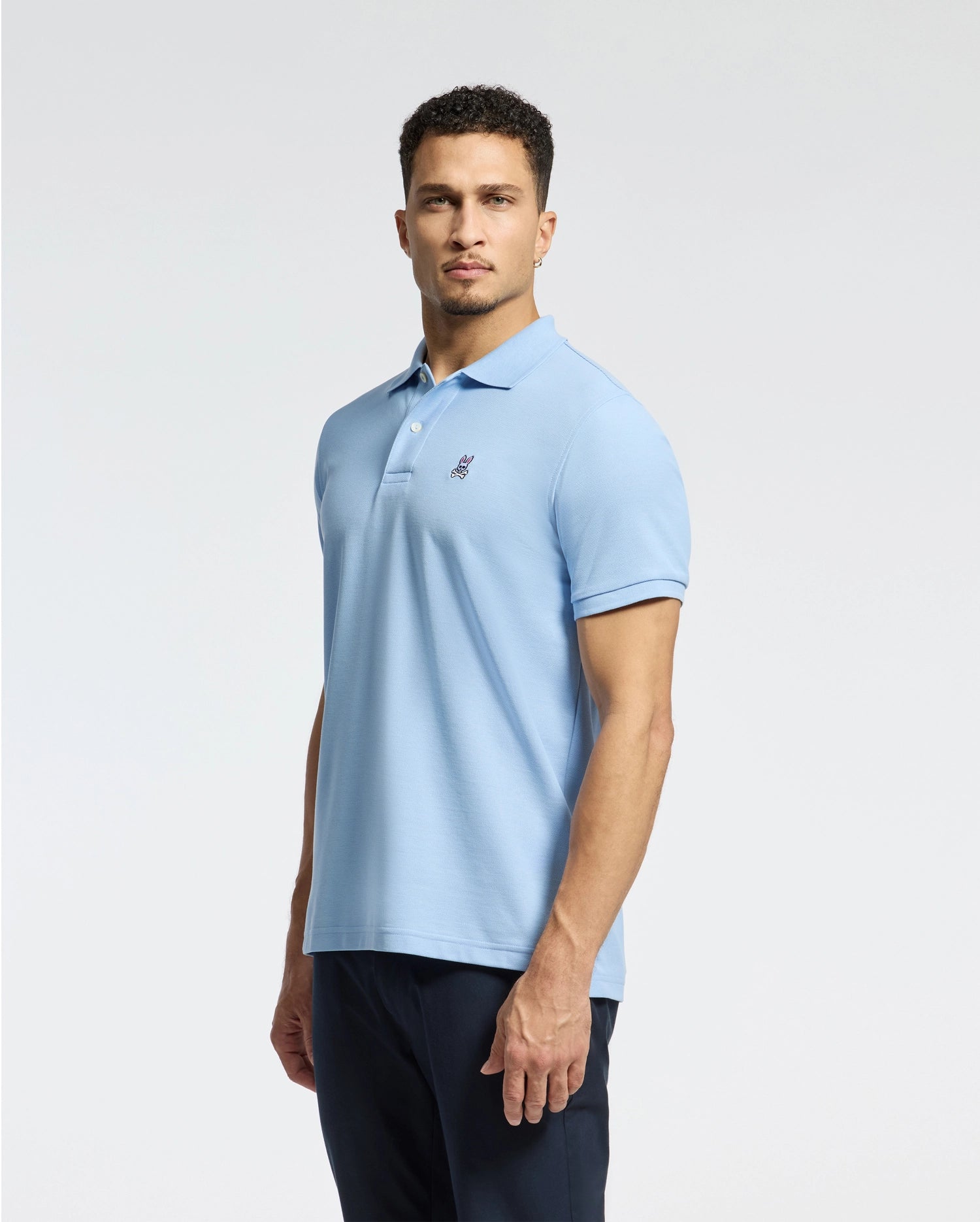 A person with short curly hair is wearing a Psycho Bunny men's classic pique polo shirt in light blue, featuring an embroidered Bunny logo on the chest and crafted from soft Pima cotton. They pair it with dark pants against a plain white background.