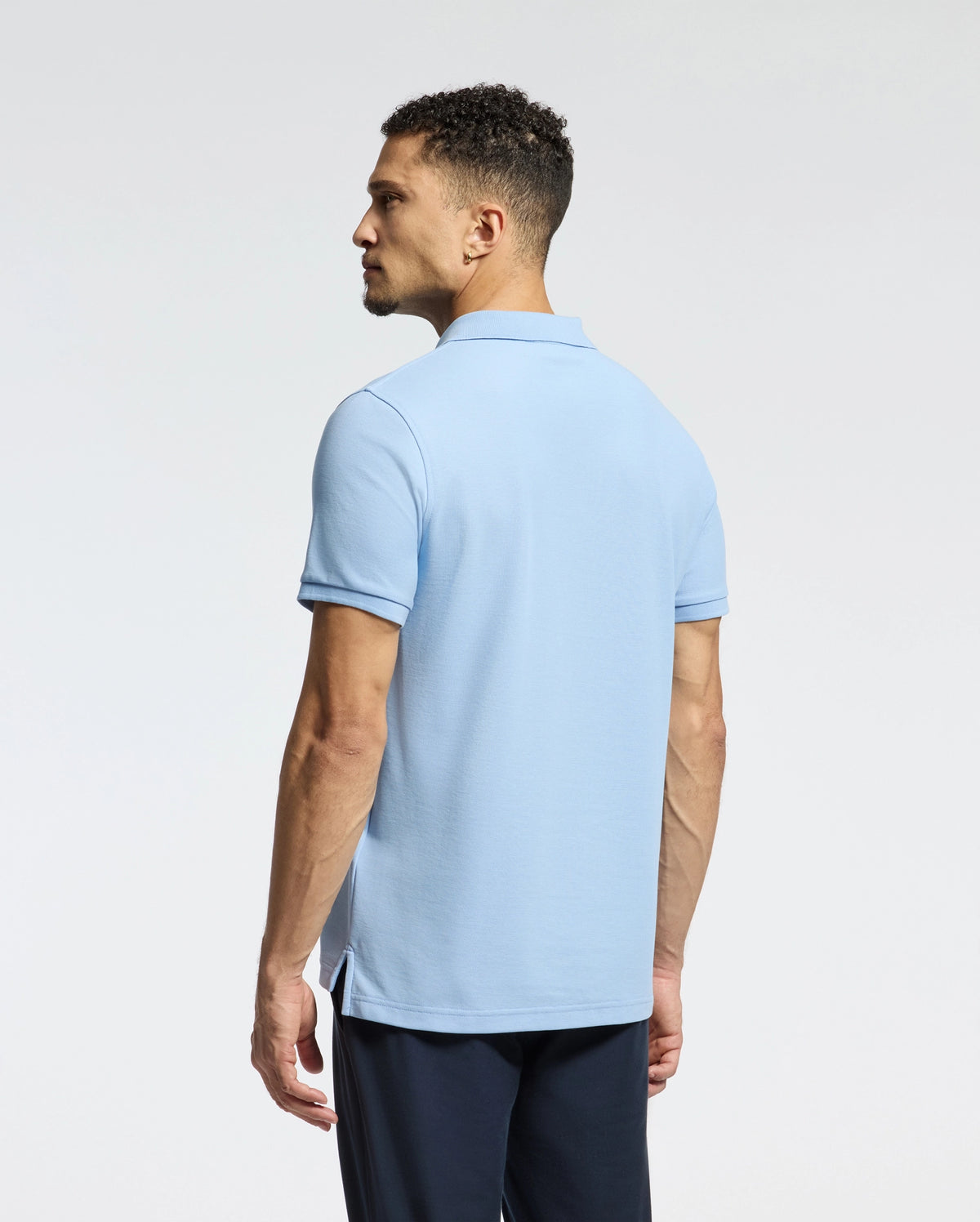 A man with short curly hair, viewed from behind, wears a light blue Psycho Bunny MENS CLASSIC PIQUE POLO SHIRT - B6K001B200 made of soft Pima cotton. He completes the look with dark pants against a white background, featuring an embroidered Bunny logo on his shirt.