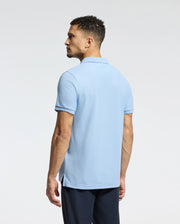 A man with short curly hair, viewed from behind, wears a light blue Psycho Bunny MENS CLASSIC PIQUE POLO SHIRT - B6K001B200 made of soft Pima cotton. He completes the look with dark pants against a white background, featuring an embroidered Bunny logo on his shirt.