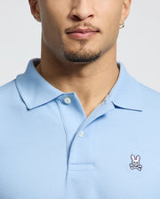 A person wears a Psycho Bunny MENS CLASSIC PIQUE POLO SHIRT - B6K001B200, made from soft Pima cotton with an embroidered logo featuring a white skull and crossbones with bunny ears. The person's face is partially visible, displaying a beard and hoop earring.