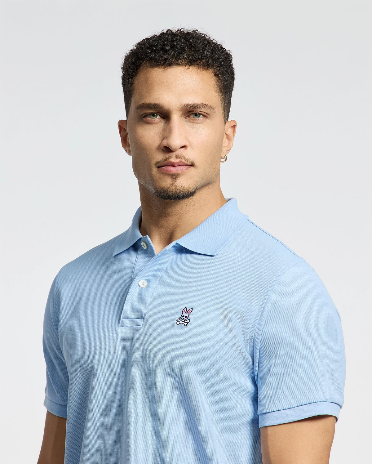 A person with short curly hair is wearing a Psycho Bunny Men's Classic Pique Polo Shirt (B6K001B200) in light blue, featuring an embroidered pink and white Bunny logo, against a plain light-colored background.