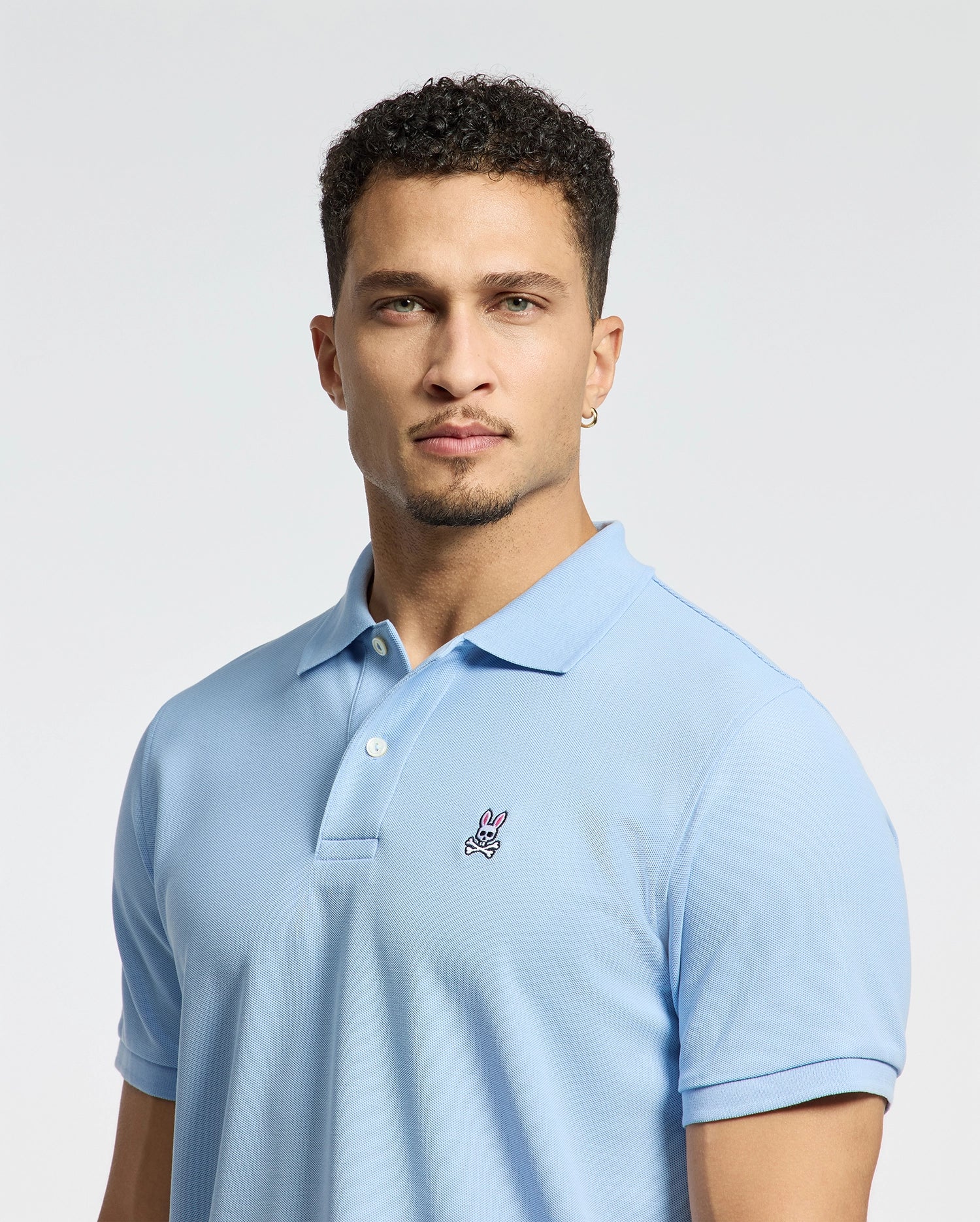 A person with short curly hair is wearing a Psycho Bunny Men's Classic Pique Polo Shirt (B6K001B200) in light blue, featuring an embroidered pink and white Bunny logo, against a plain light-colored background.