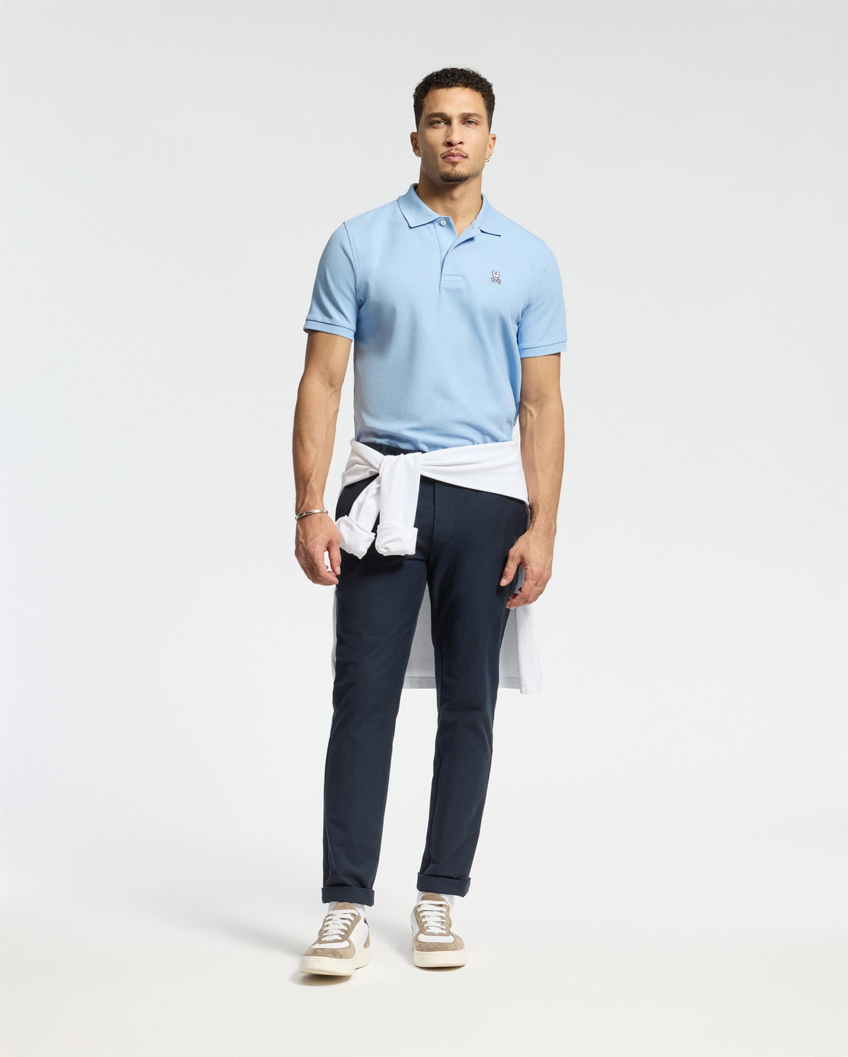 A man with short hair and a neutral expression stands against a plain backdrop wearing dark blue pants, brown sneakers, and a light blue Psycho Bunny MENS CLASSIC PIQUE POLO SHIRT - B6K001B200 with an embroidered Bunny logo. A white sweater is tied around his waist.