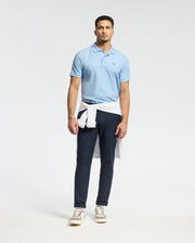 A man with short hair and a neutral expression stands against a plain backdrop wearing dark blue pants, brown sneakers, and a light blue Psycho Bunny MENS CLASSIC PIQUE POLO SHIRT - B6K001B200 with an embroidered Bunny logo. A white sweater is tied around his waist.