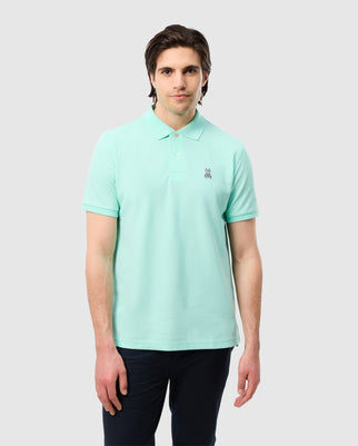 A person in a light green Psycho Bunny Men's Classic Pique Polo Shirt, B6K001C200, made of soft Pima cotton with a small chest logo, pairs it with dark pants and stands against a plain gray background, looking neutrally at the camera.