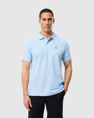 A person in a Psycho Bunny Men's Classic Pique Polo, light blue with an embroidered logo and genuine Mother of Pearl buttons, stands against a neutral background. They have short dark hair and wear dark pants.