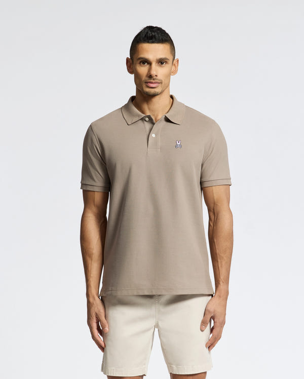 A man stands facing forward in a light brown Psycho Bunny MENS CLASSIC PIQUE POLO SHIRT (B6K001CRPC) with an embroidered chest logo, paired with beige shorts, against a plain light background.