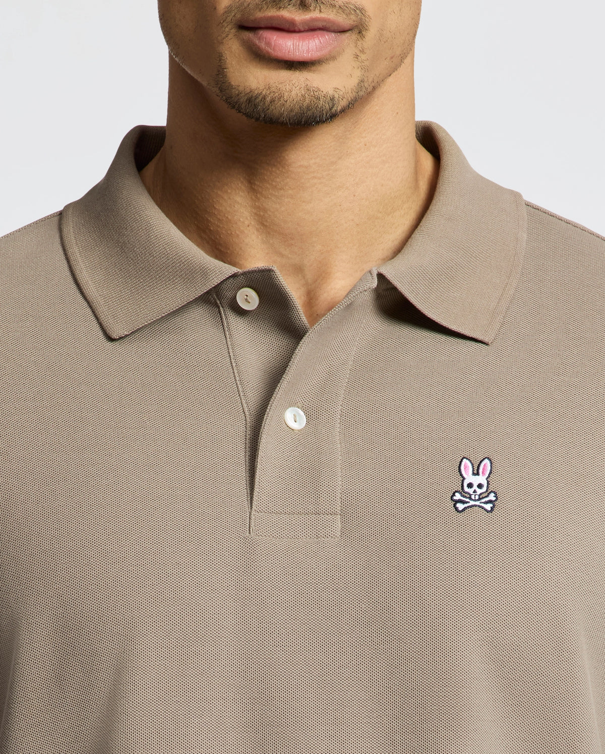 A man wears a taupe Psycho Bunny MENS CLASSIC PIQUE POLO SHIRT (B6K001CRPC) with a collar and buttons, made from soft Pima cotton. A pink bunny and white skull-and-crossbones patch is on the left chest. His face is partially visible.