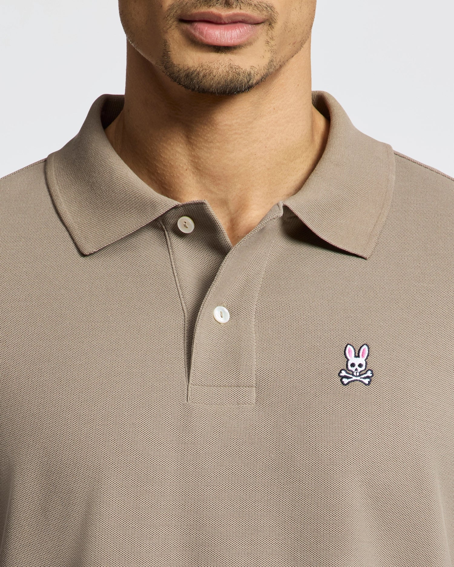 A man wears a taupe Psycho Bunny MENS CLASSIC PIQUE POLO SHIRT (B6K001CRPC) with a collar and buttons, made from soft Pima cotton. A pink bunny and white skull-and-crossbones patch is on the left chest. His face is partially visible.