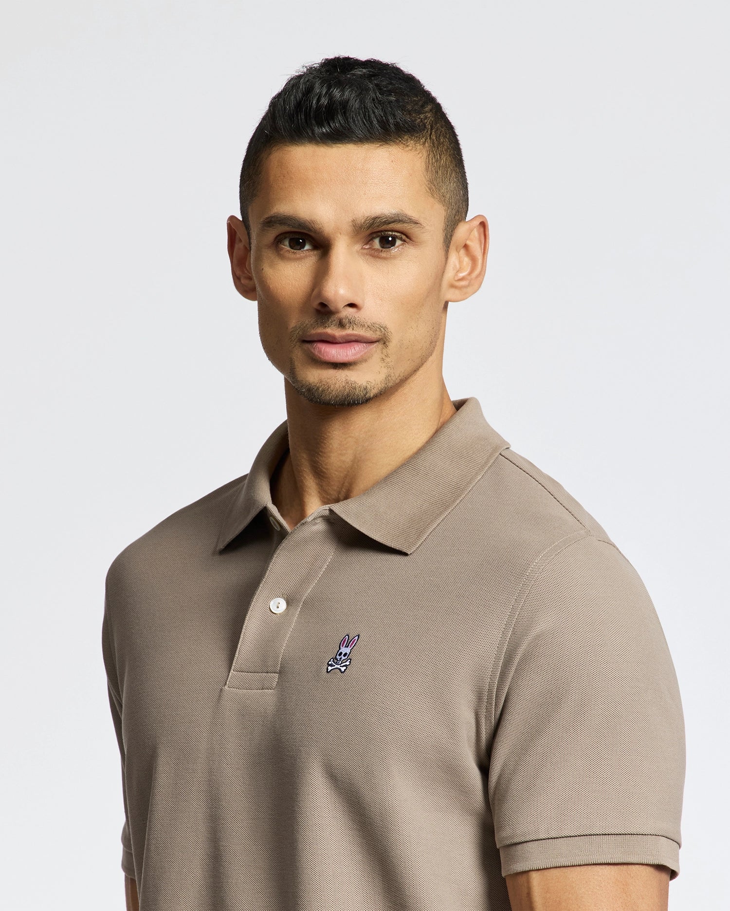 A man with short dark hair and light facial hair wears a Psycho Bunny MENS CLASSIC PIQUE POLO SHIRT - B6K001CRPC in taupe, featuring a small embroidered bunny head on the chest, standing against a plain light background.