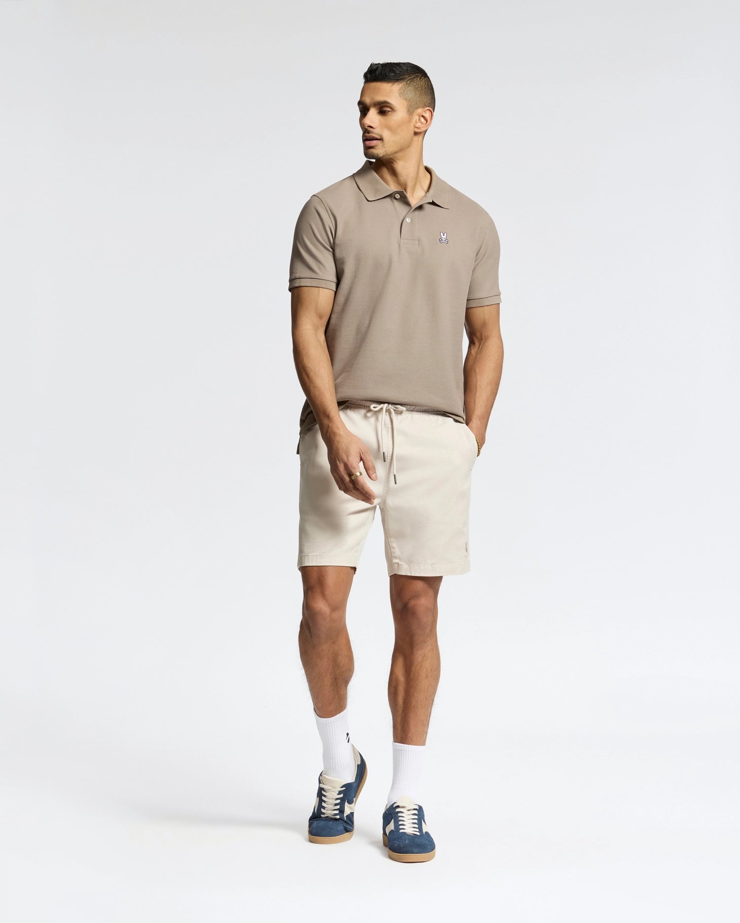 A man stands against a white background wearing a Psycho Bunny MENS CLASSIC PIQUE POLO SHIRT (B6K001CRPC) in beige, paired with light beige shorts, white socks, and navy sneakers. His hands are in his pockets as he looks to the side.