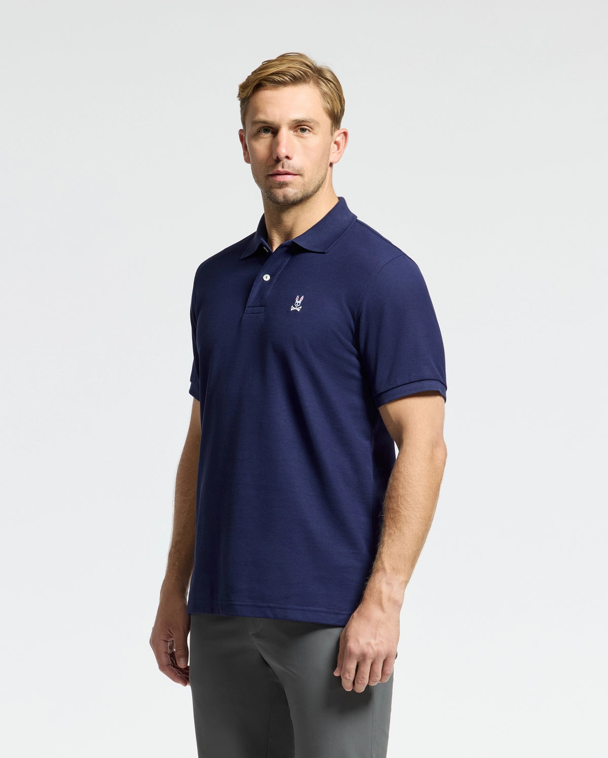 A man wearing the Psycho Bunny Men's Classic Pique Polo Shirt in a regular fit navy blue, made from soft pima cotton and featuring a small chest logo, stands against a plain background.
