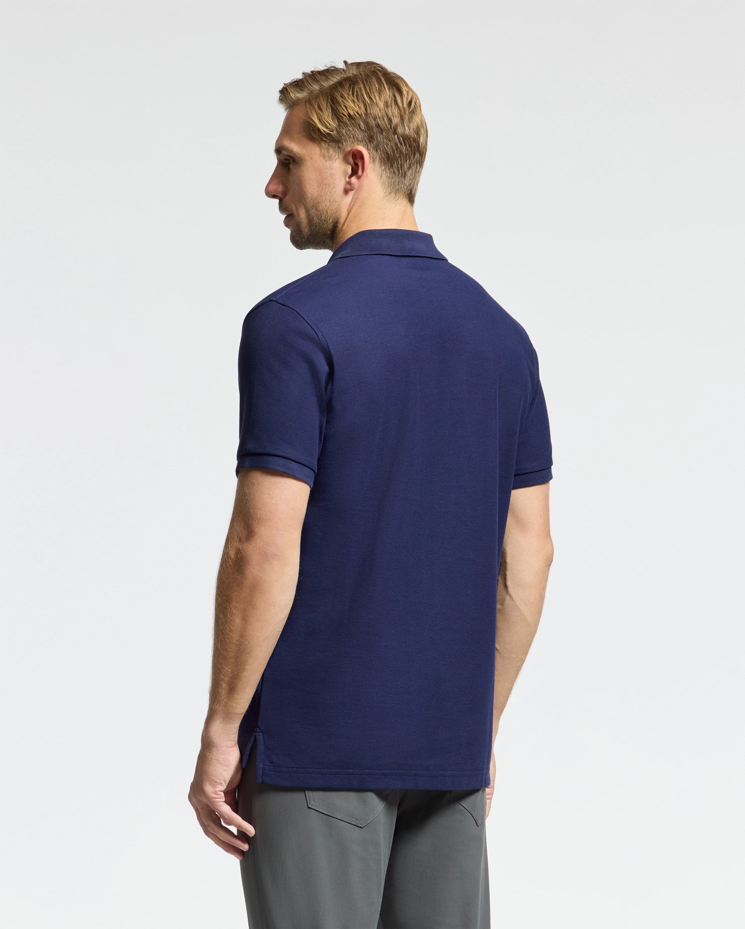 A man with light brown hair is shown from behind, wearing a Psycho Bunny navy blue Men's Classic Pique Polo Shirt (Model: B6K001CRPC) made from pima cotton and gray pants. He stands against a simple, light gray background.