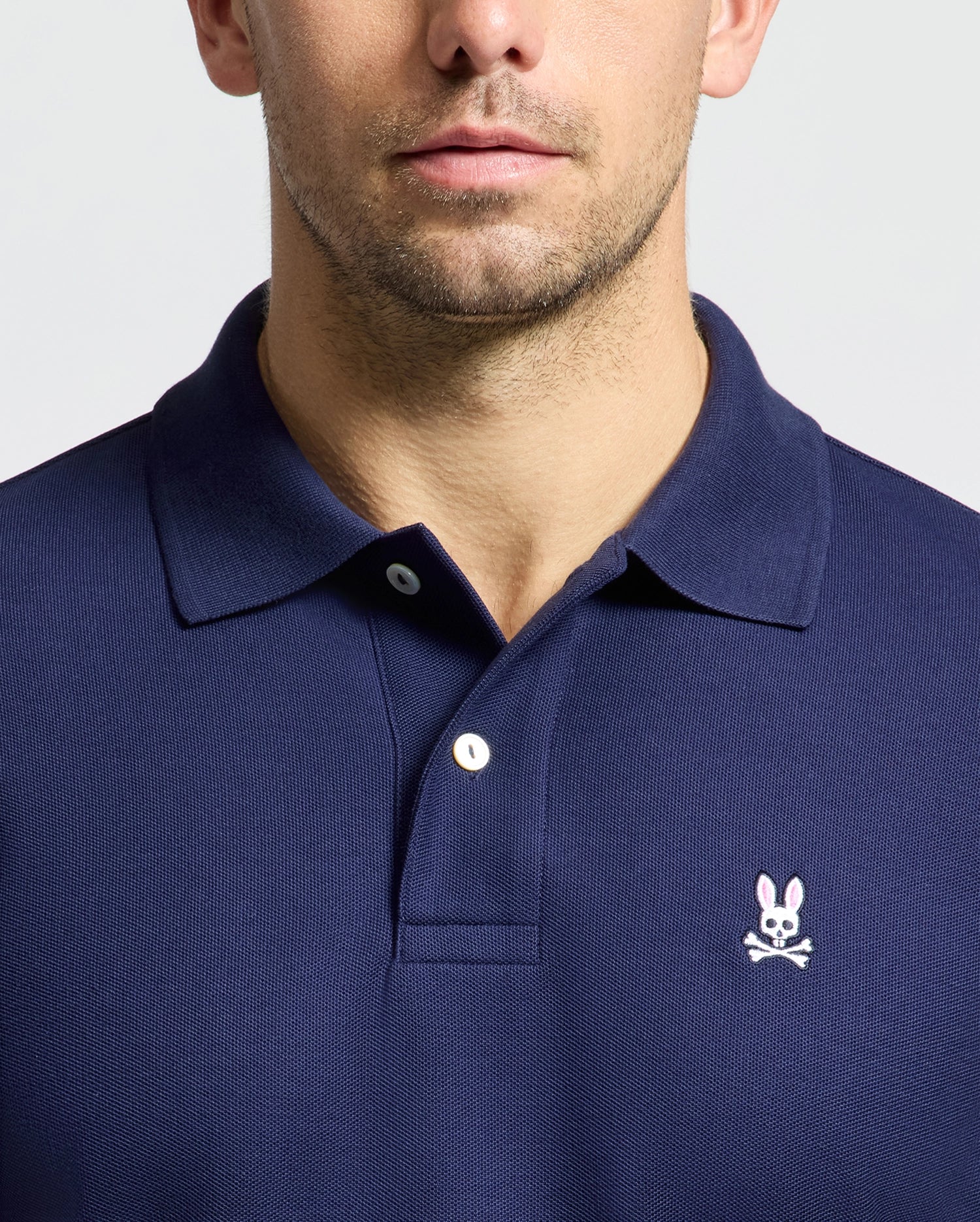 A man is dressed in a regular fit, navy blue polo shirt made from soft pima cotton, displaying the pink Psycho Bunny logo with crossbones on the chest. The backdrop is simple and light-toned.