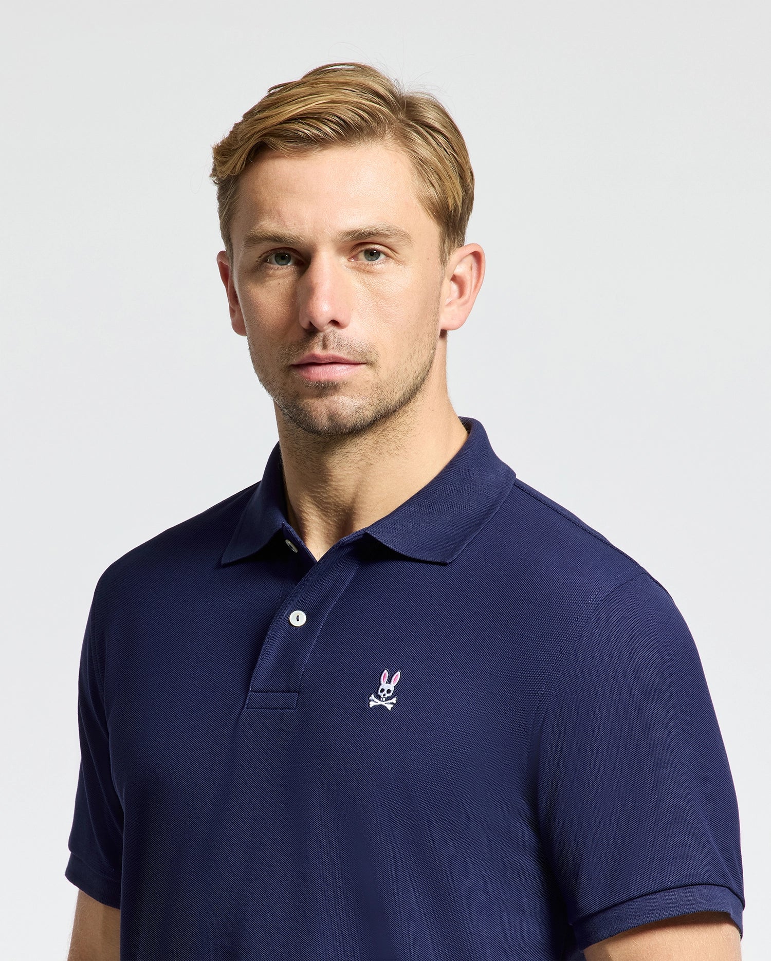 A man with short blonde hair is wearing the Psycho Bunny Men's Classic Pique Polo Shirt - B6K001CRPC in navy blue, which features a small embroidered rabbit logo on the chest. Made from pima cotton for comfort, this regular fit shirt enhances his appearance as he looks straight into the camera against a simple, light background.