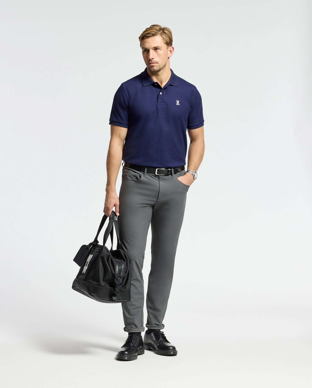 A man wearing a Psycho Bunny MENS CLASSIC PIQUE POLO SHIRT in navy and gray trousers stands against a plain backdrop. He holds a black duffel bag in one hand, complemented by black shoes and a watch.