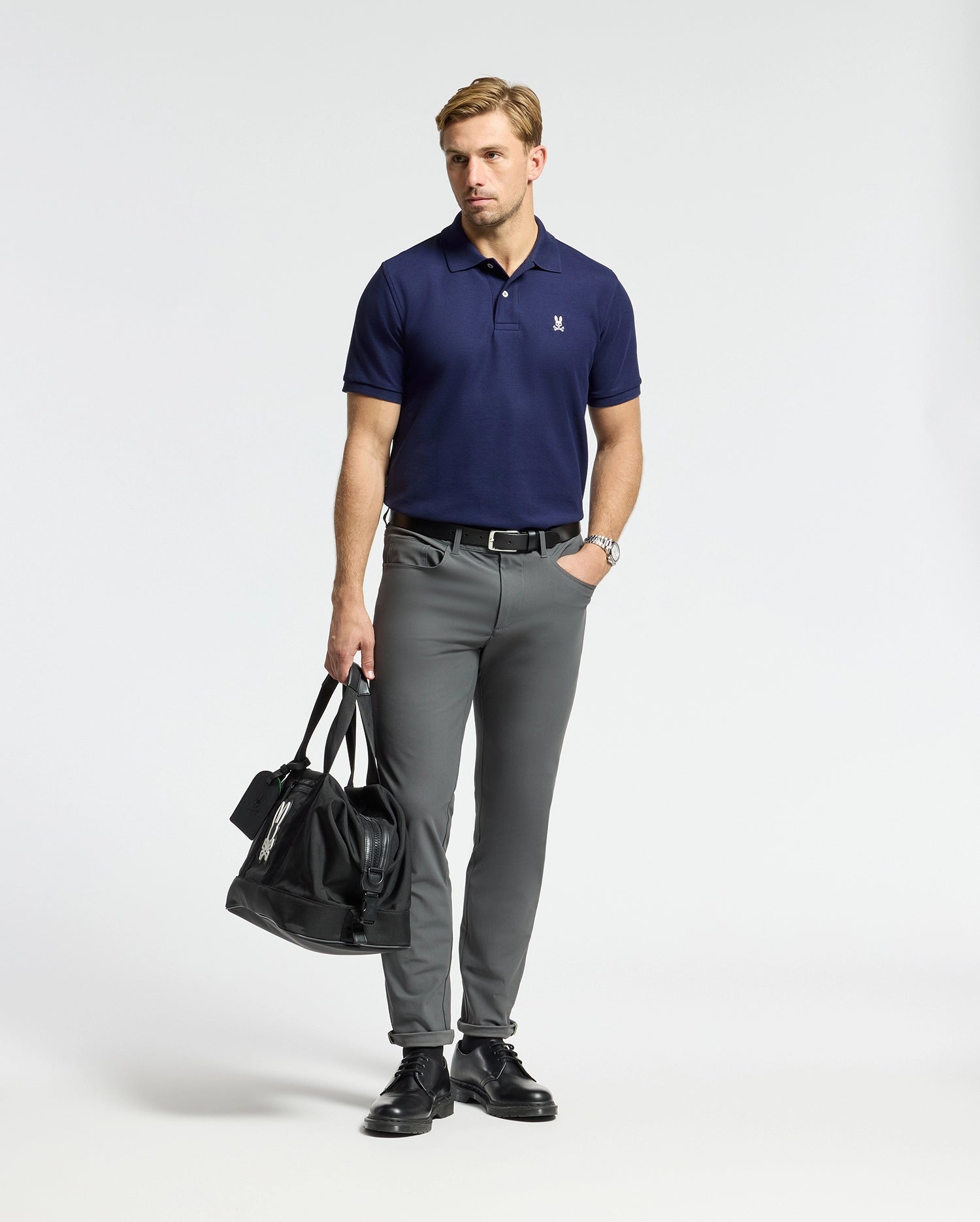 A man wearing a Psycho Bunny MENS CLASSIC PIQUE POLO SHIRT in navy and gray trousers stands against a plain backdrop. He holds a black duffel bag in one hand, complemented by black shoes and a watch.