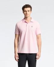A man stands against a white background, wearing Psycho Bunny's light pink Mens Classic Pique Polo Shirt (B6K001CRPC) made of Peruvian pima cotton with a small chest emblem. He has curly hair, looks directly at the camera, and is also dressed in black pants.