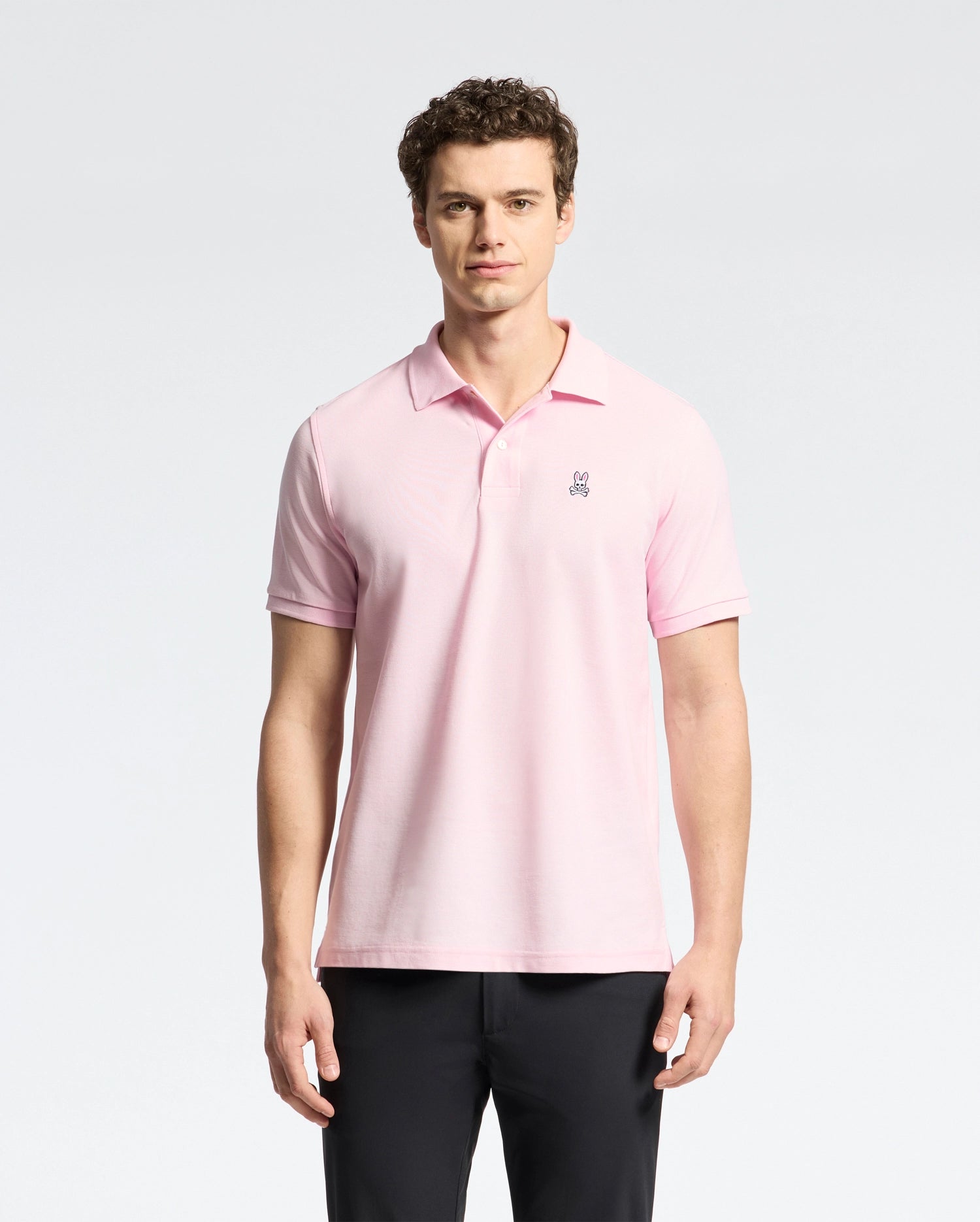 A man stands against a white background, wearing Psycho Bunny's light pink Mens Classic Pique Polo Shirt (B6K001CRPC) made of Peruvian pima cotton with a small chest emblem. He has curly hair, looks directly at the camera, and is also dressed in black pants.