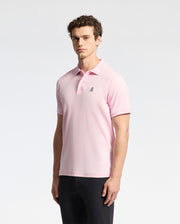 A man wears a Psycho Bunny MENS CLASSIC PIQUE POLO SHIRT (B6K001CRPC) in light pink Peruvian pima cotton, with an embroidered logo on the chest, paired with black pants against a plain white background.