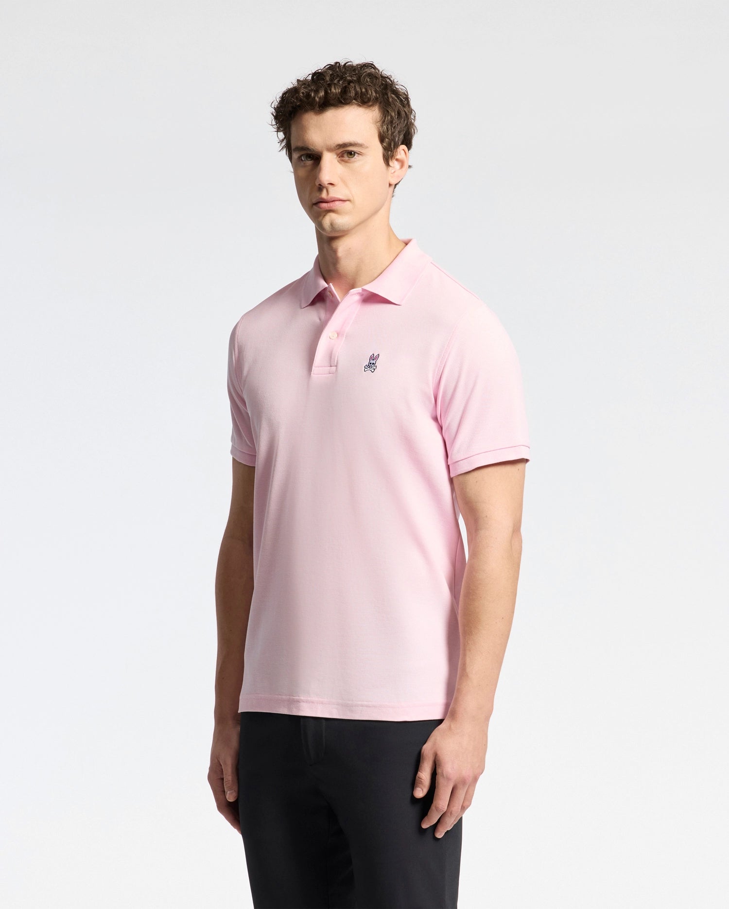 A man wears a Psycho Bunny MENS CLASSIC PIQUE POLO SHIRT (B6K001CRPC) in light pink Peruvian pima cotton, with an embroidered logo on the chest, paired with black pants against a plain white background.