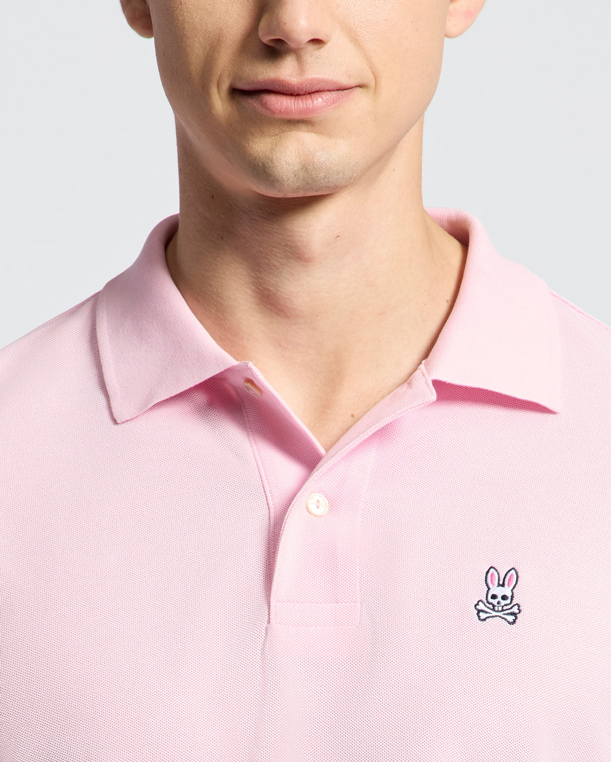 A person is wearing the Psycho Bunny MENS CLASSIC PIQUE POLO SHIRT - B6K001CRPC in pink, featuring a bunny and crossbones logo on the left chest. The photo highlights their upper chest and partially visible face against a simple light background.