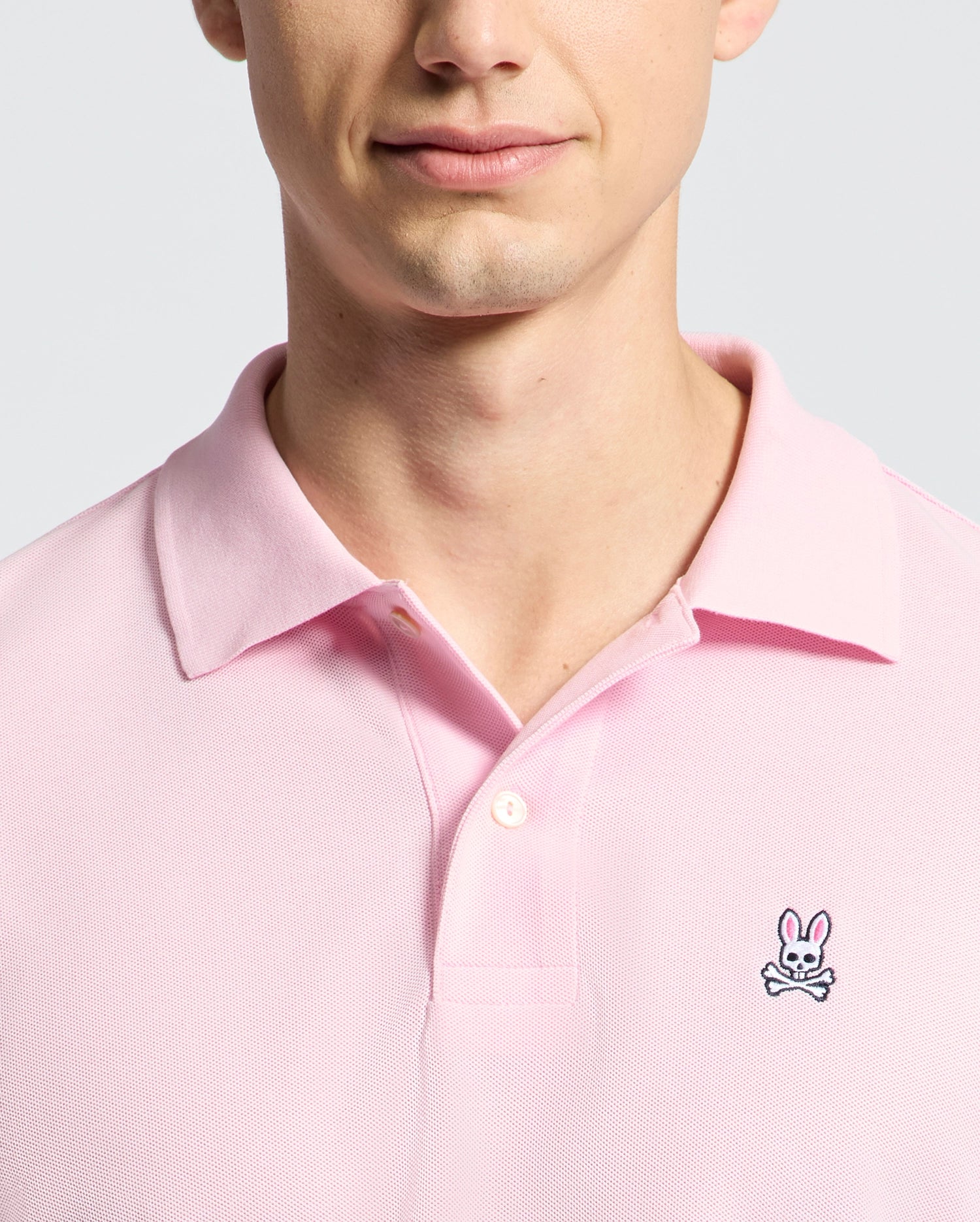 A person is wearing the Psycho Bunny MENS CLASSIC PIQUE POLO SHIRT - B6K001CRPC in pink, featuring a bunny and crossbones logo on the left chest. The photo highlights their upper chest and partially visible face against a simple light background.