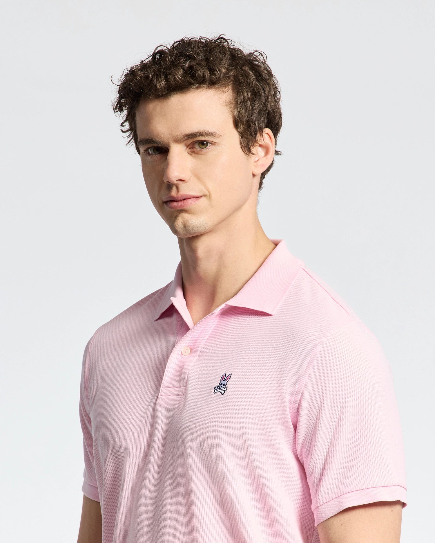A person with curly hair wears a Psycho Bunny MENS CLASSIC PIQUE POLO SHIRT - B6K001CRPC in light pink, made of luxurious Peruvian pima cotton, featuring a small embroidered bunny logo. They stand against a plain gray background, looking slightly to the side.