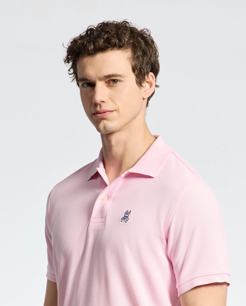 A person with curly hair wears a Psycho Bunny MENS CLASSIC PIQUE POLO SHIRT - B6K001CRPC in light pink, made of luxurious Peruvian pima cotton, featuring a small embroidered bunny logo. They stand against a plain gray background, looking slightly to the side.