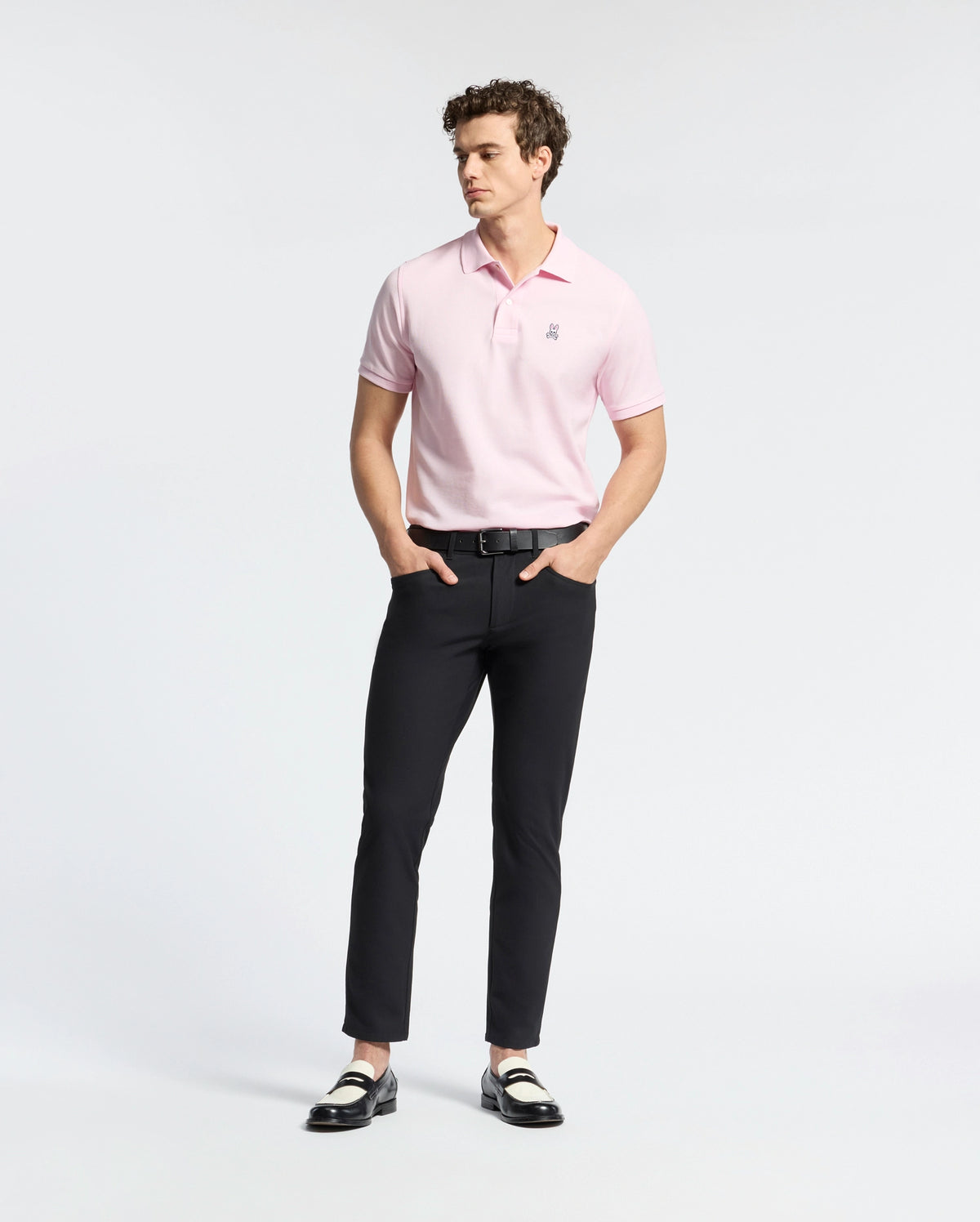 A person wearing a light pink Psycho Bunny MENS CLASSIC PIQUE POLO SHIRT - B6K001CRPC, black pants, and black and white loafers stands against a plain white background. They have curly hair and their hands are in their pockets.