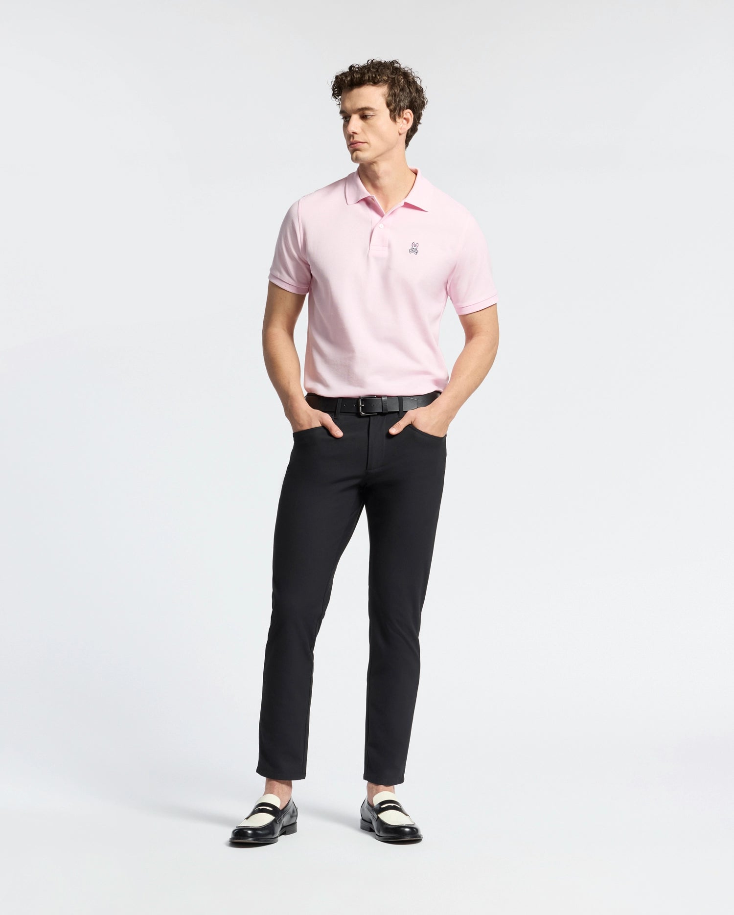 A person wearing a light pink Psycho Bunny MENS CLASSIC PIQUE POLO SHIRT - B6K001CRPC, black pants, and black and white loafers stands against a plain white background. They have curly hair and their hands are in their pockets.