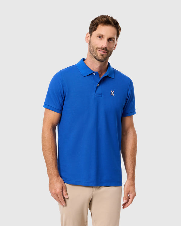 A man with short hair and a beard models the bright blue Men's Classic Pique Polo Shirt by Psycho Bunny (B6K001CRPC) against a plain white backdrop. Made from luxurious Pima cotton, it showcases an embroidered logo on the chest, enhancing his sophisticated appearance.
