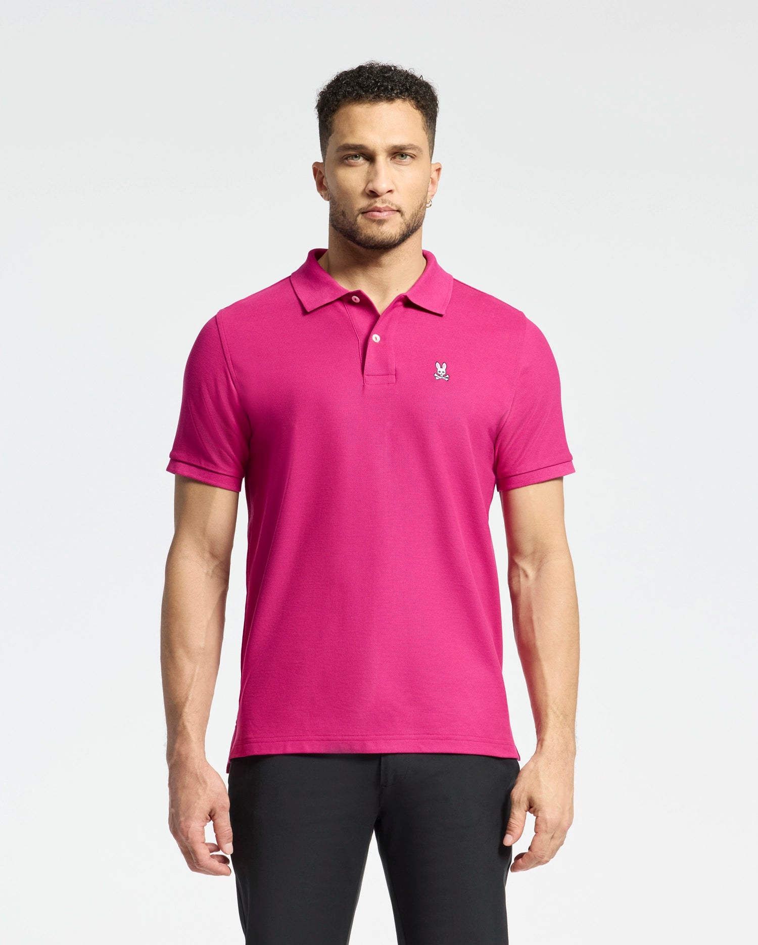 A man stands against a light background wearing the Psycho Bunny MENS CLASSIC PIQUE POLO SHIRT - B6K001CRPC in bright pink Pima cotton, featuring an embroidered Bunny logo on the chest. He's paired it with dark pants, and his short curly hair highlights his neutral expression.