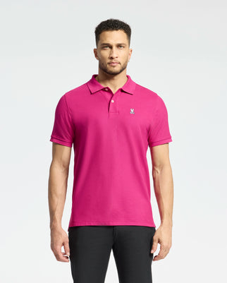 A man stands against a light background wearing the Psycho Bunny MENS CLASSIC PIQUE POLO SHIRT - B6K001CRPC in bright pink Pima cotton, featuring an embroidered Bunny logo on the chest. He's paired it with dark pants, and his short curly hair highlights his neutral expression.