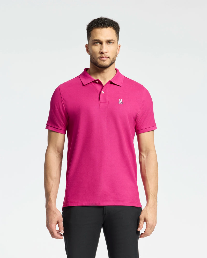 A man stands against a light background wearing the Psycho Bunny MENS CLASSIC PIQUE POLO SHIRT - B6K001CRPC in bright pink Pima cotton, featuring an embroidered Bunny logo on the chest. He's paired it with dark pants, and his short curly hair highlights his neutral expression.